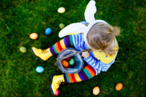 Close-up of legs of toddler girl with colorful stockings and shoes and basket with colored eggs. Child having fun with traditional Easter eggs hunt, outdoors. Unrecognizable face, no face