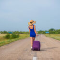 A woman in a hat and with a big bag is walking along the roadway. Girl in a blue tight-fitting dress with a purple suitcase on the track. Brunette with a sports figure on the road