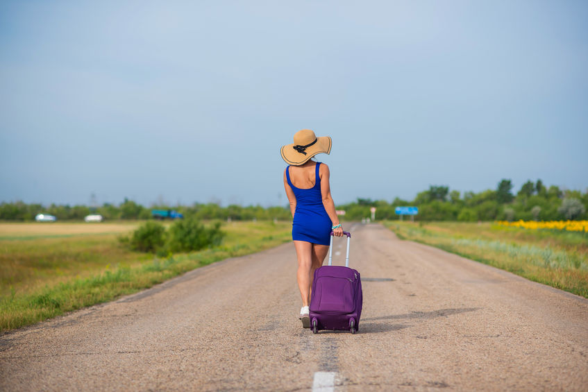 A woman in a hat and with a big bag is walking along the roadway. Girl in a blue tight-fitting dress with a purple suitcase on the track. Brunette with a sports figure on the road